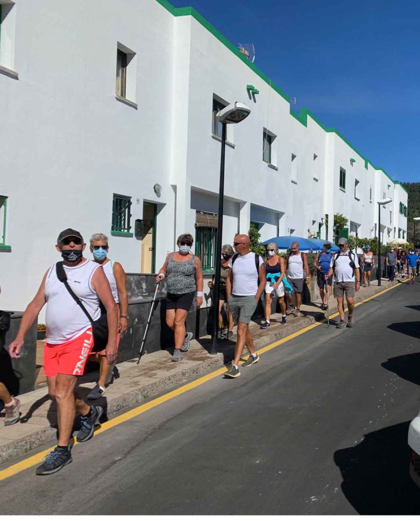 Menschen in sommerlicher Kleidung, kurzen Hosen laufen auf einer kleinen Strasse hintereinander. Sie tragen Mundschutzmasken wegen CoVid.