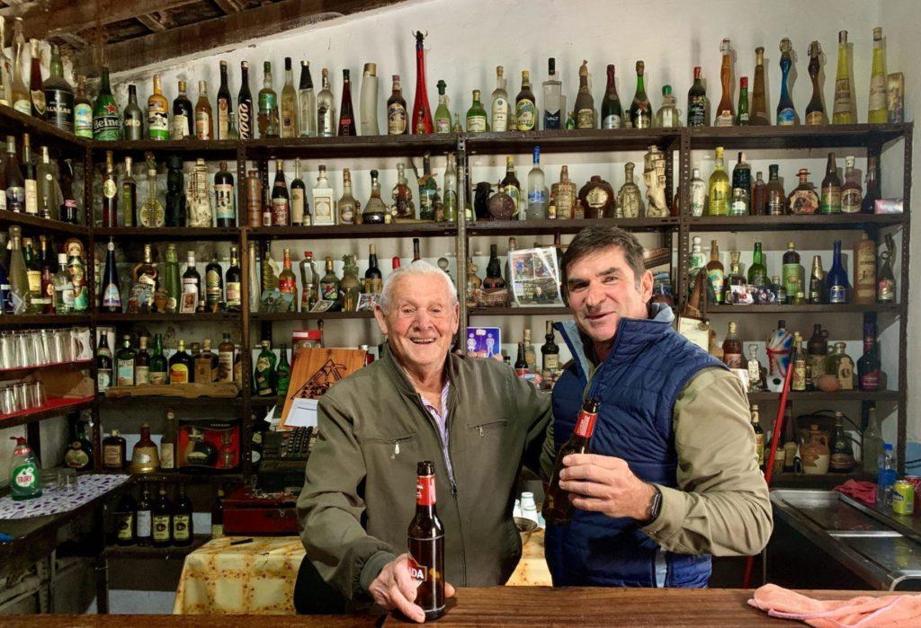 zwei ältere Herren stehen hinter einer Theke jeder hält eine Bierflasche in der Hand. Im Hintergrund ein großes Regal mit vielen Flaschen Spirituosen