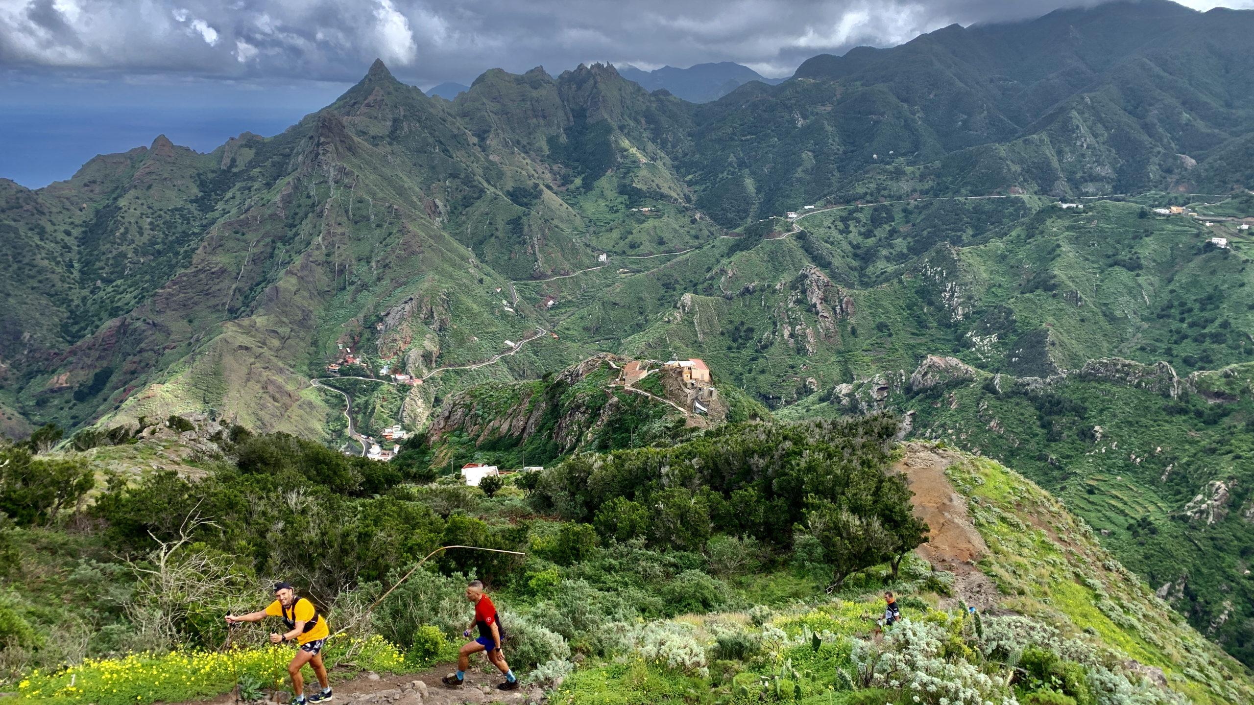 zwei sportliche Wanderer in bunter Kleidung gehen einen Bergpfad hinauf. Im Hintergrund das zerklüftete Bergmassiv des Anaga Gebirges auf Teneriffa