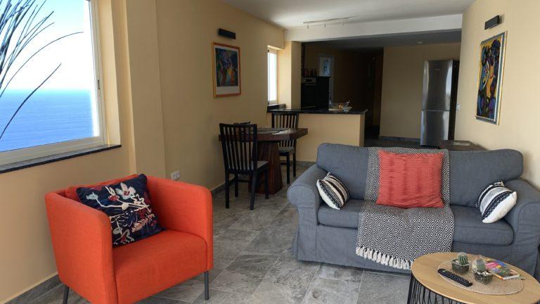 Blick in ein Wohnzimmer mit grauem Sofa, rotem Sessel, Holz Esstisch mit 2 Stühlen mit