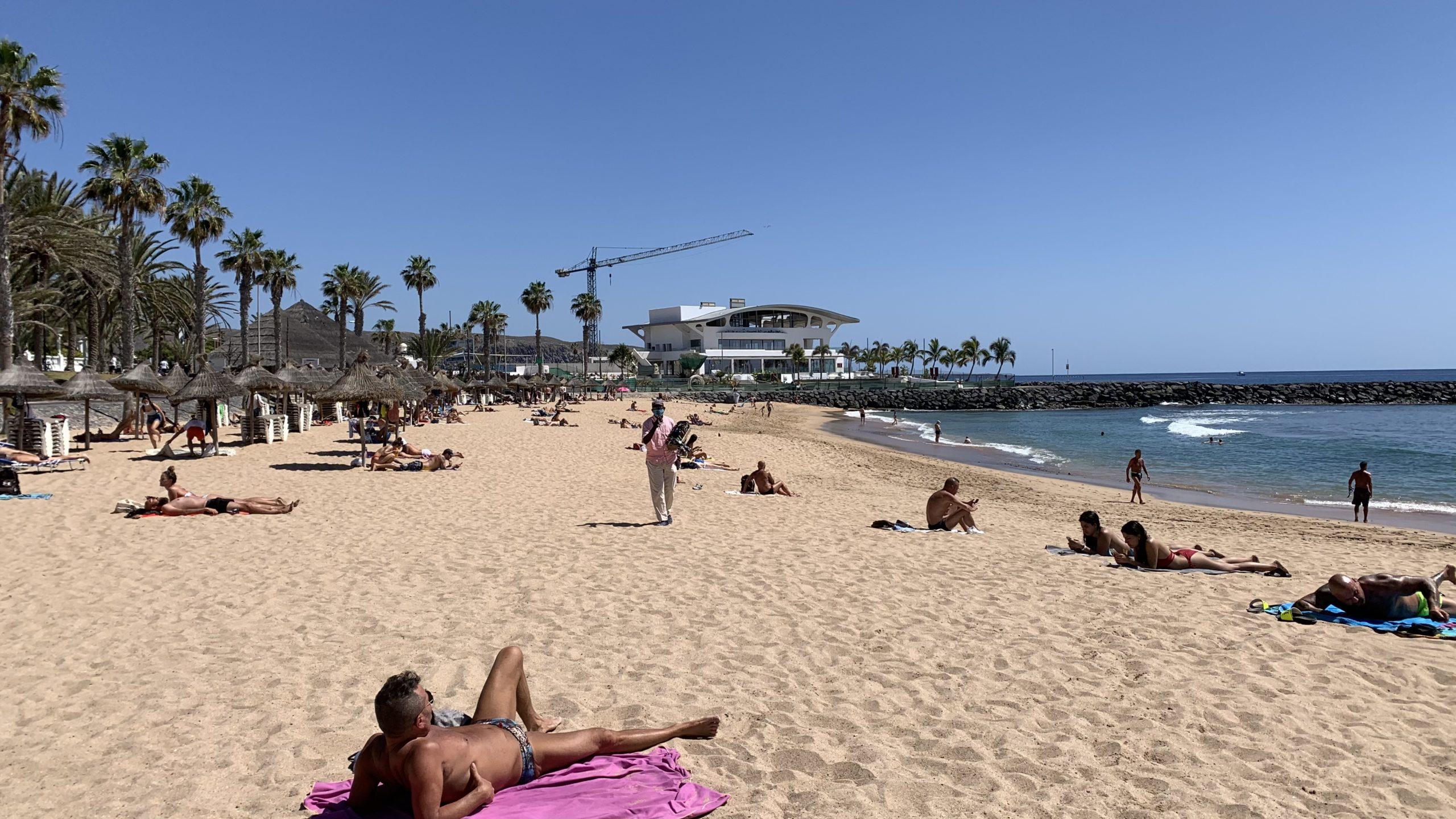 Mehrere Touristen liegen an einem Sandstrand bei Sonnenschein und blauem Wasser